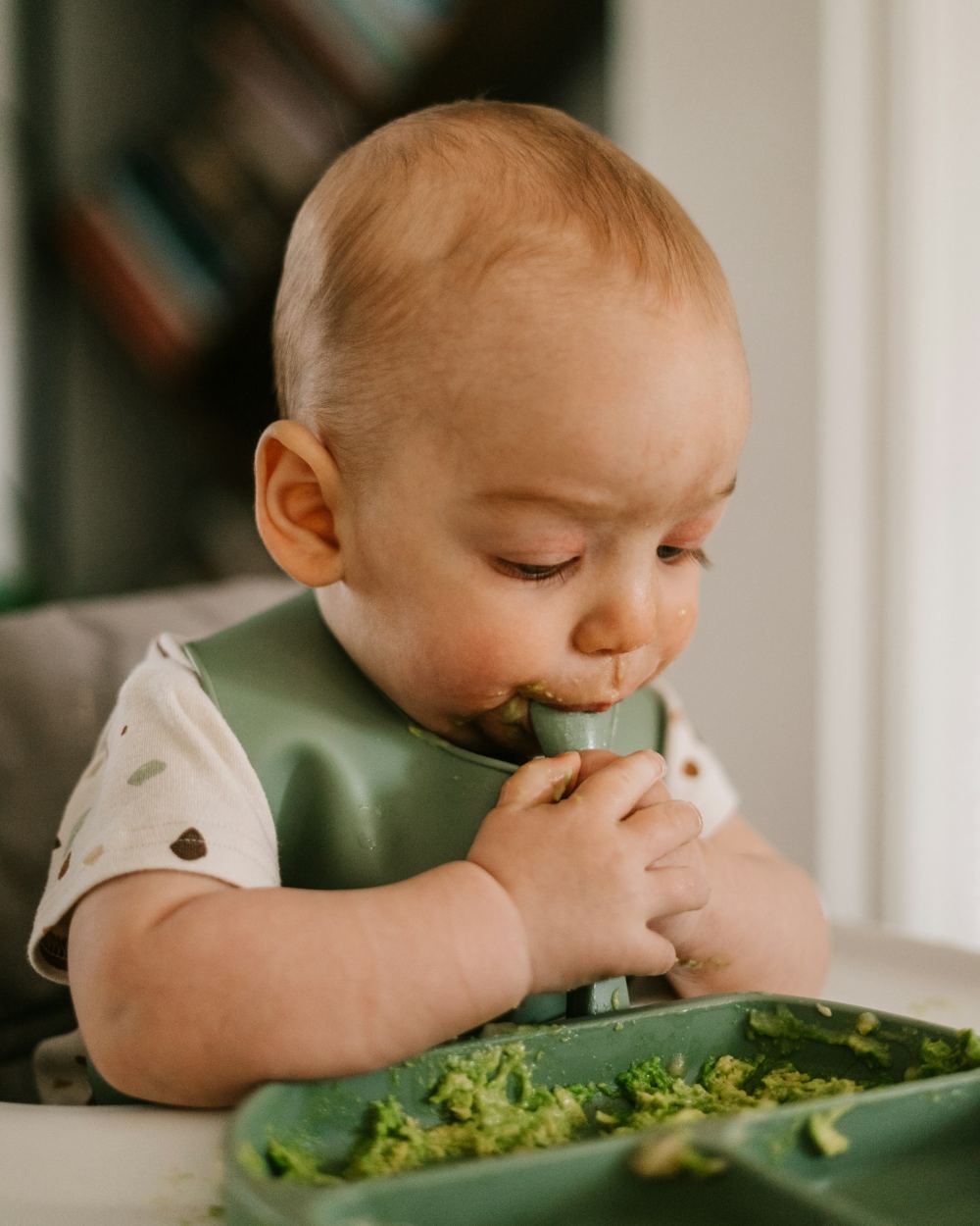 Un bambino sul seggiolone mangia da solo una purea di verdure, con il bavaglino e il cucchiaino in mano